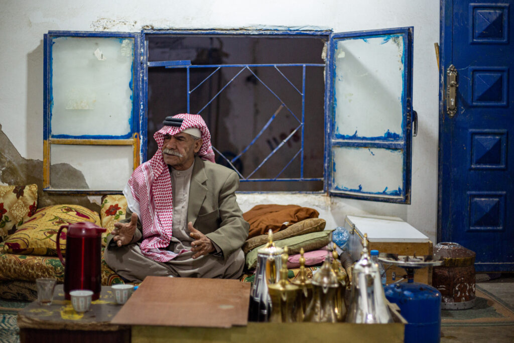 A man dressed in a red and white keffiyeh and tan suit sits on a sofa covered in pillows and gestures with his hands.