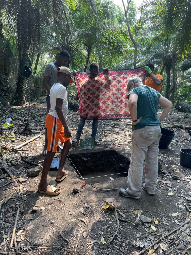 Two people hold a decorated red and beige cloth over a square hole in the ground while peering into it. Three others, one of them with gray hair and lighter skin than the rest, look down into the hole.