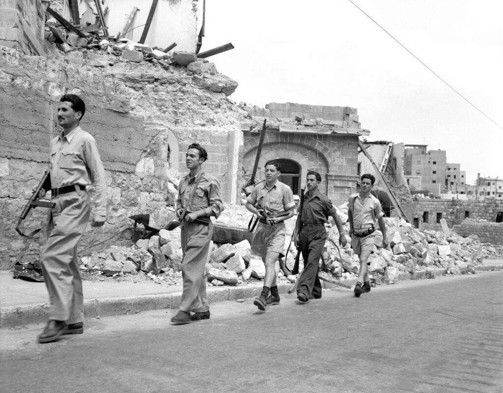 A black-and-white photograph shows a group of people walking in a line past collapsed brick buildings and piles of rubble.