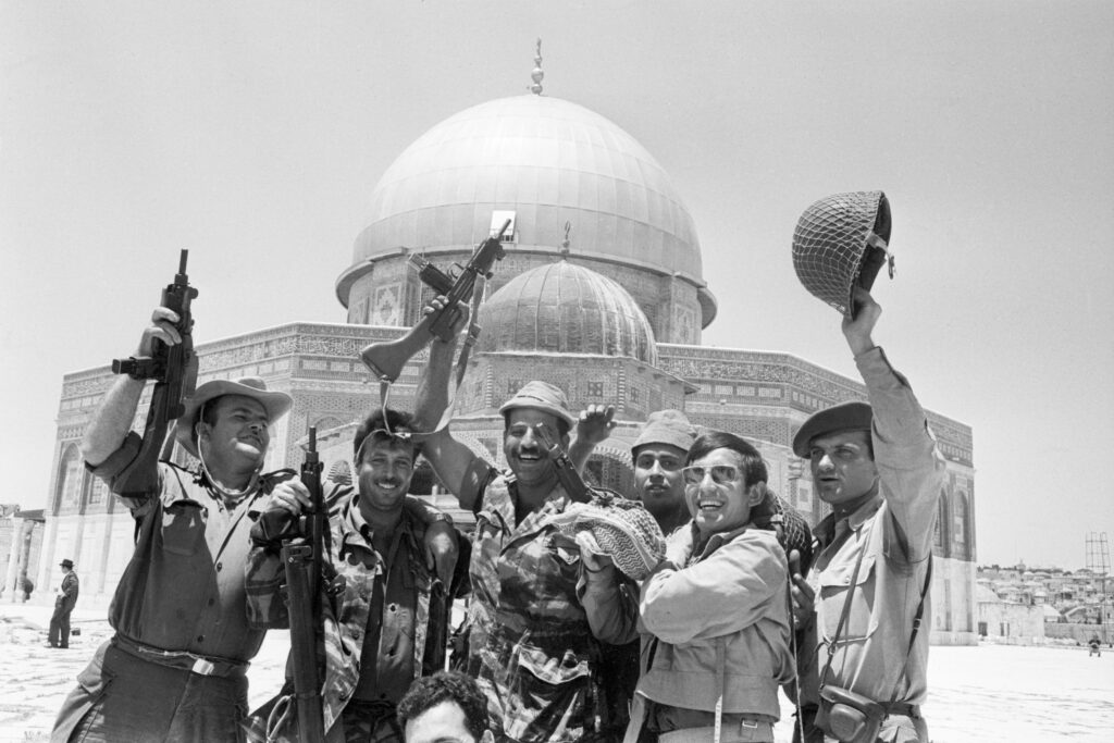 A black-and-white photograph features a group of people, with several holding up large guns, smiling and posing in front of a large, domed building.
