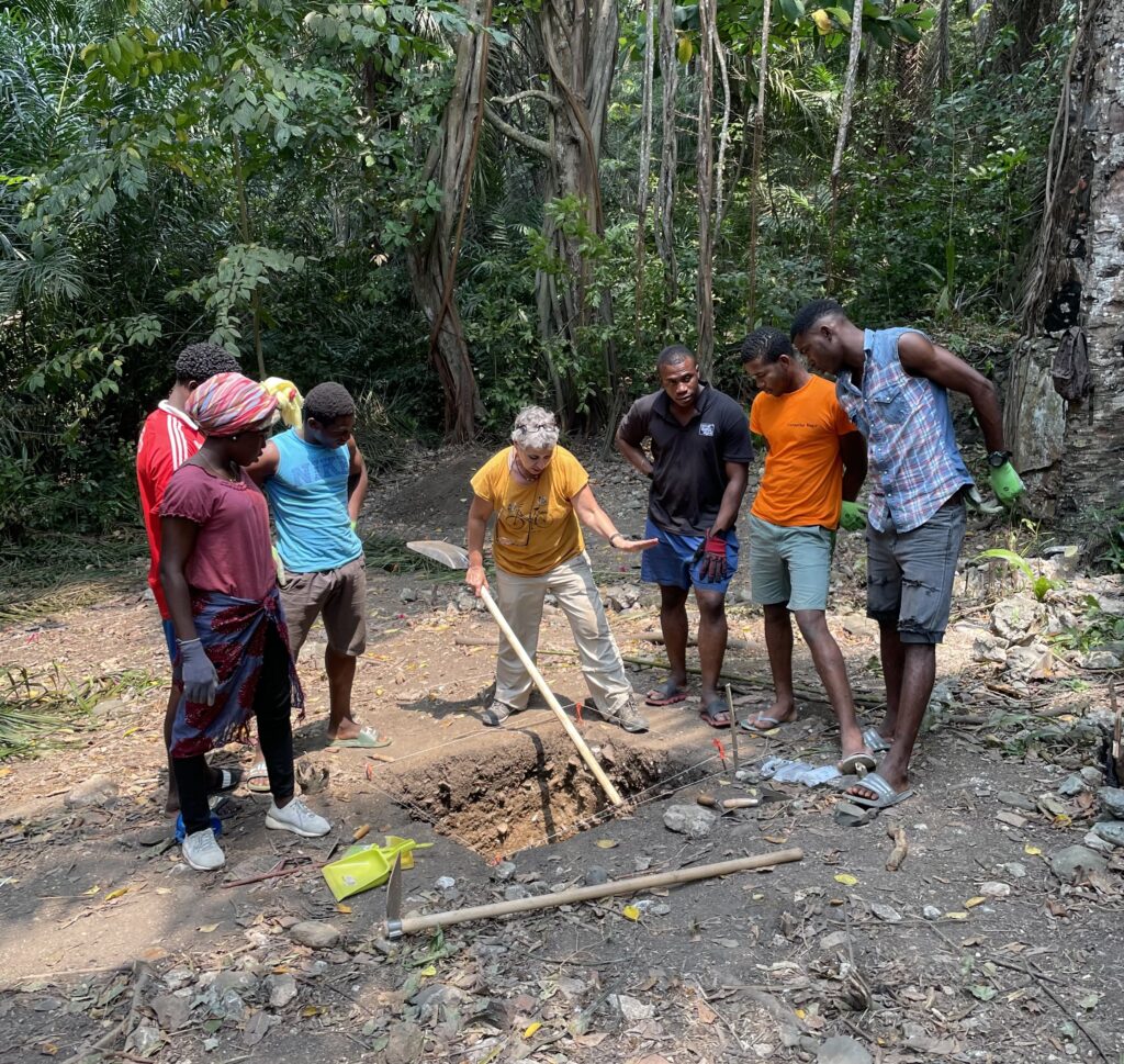 A group of people stand on an emptied dirt plot around a square hole. The one in the center, with lighter skin than the rest, holds the end of a shovel in the hole.
