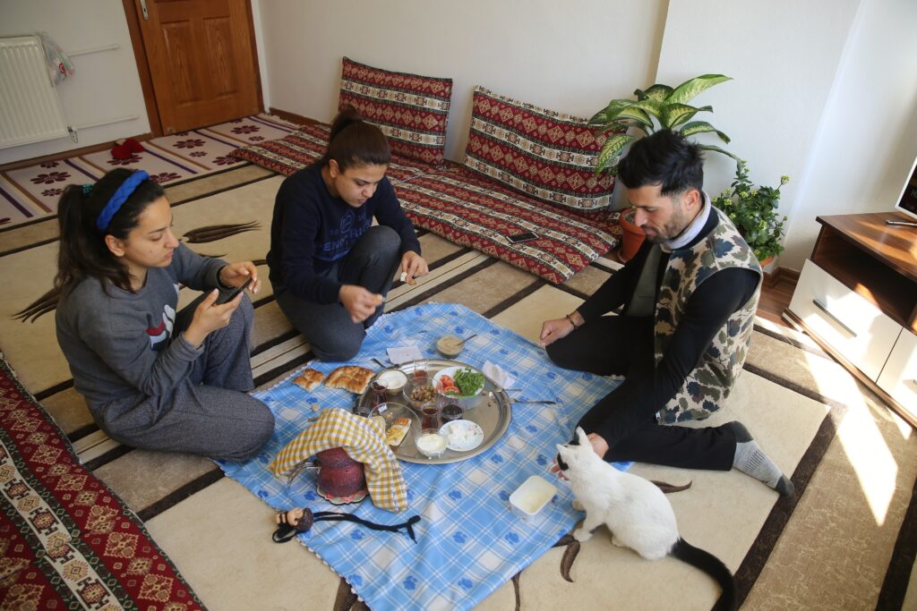 In a room with decorative auburn and white pillows lining its walls, three people sit on the floor eating from a silver tray on a blue cloth. A white and black cat sits beside the person on the right.