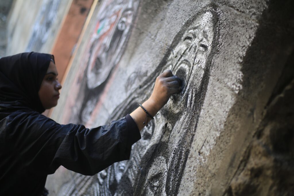 A person wearing a black headscarf and robes uses charcoal to draw an image of a screaming face on a stone wall.