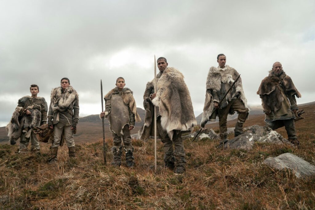 A group of people wearing large fur pelts and holding spears stand in a row on a rocky field of yellowing grasses.