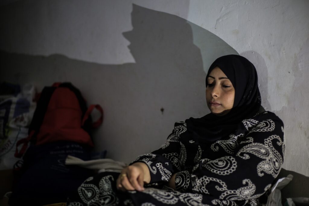 In front of a white wall with shadows cast on it, a person wearing a black headscarf and black-and-white paisley top looks down while sitting on a gray couch.