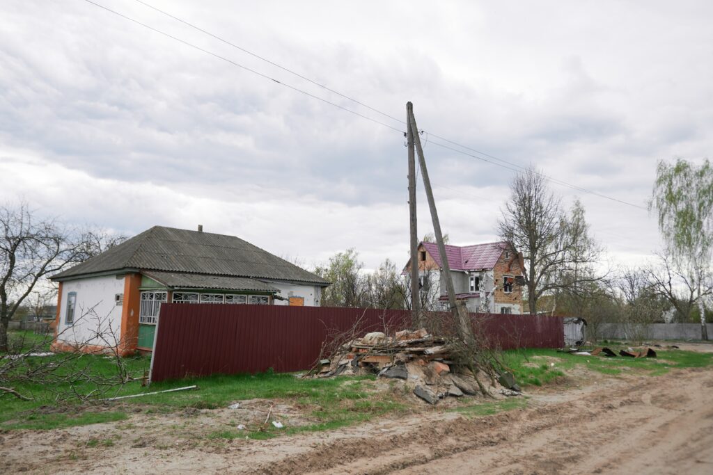 A pile of rocks sits in front of a brown fence on the side of a dirt road. A few houses line the other side of the fence.