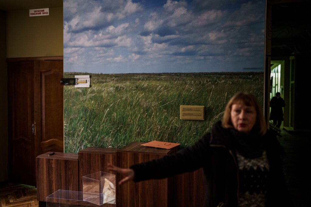 A person wearing a black shirt and jacket stands in front of a wall painted to look like a grassy field beneath a blue sky. Several wooden boxes line the wall, and clear acrylic boxes sit beside them on the floor.