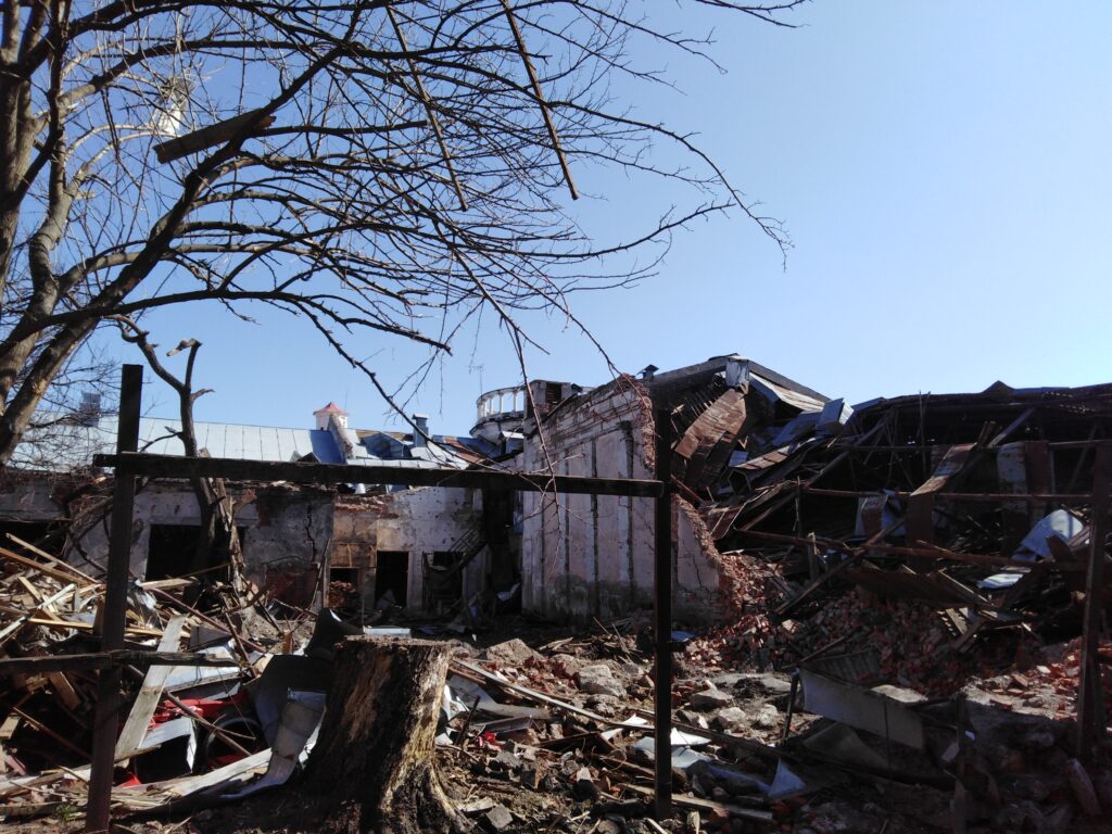 A partially destroyed L-shaped white building is shown in front of a pale blue sky. Rocks, a large tree stump, and other debris lie on the ground in front of it.