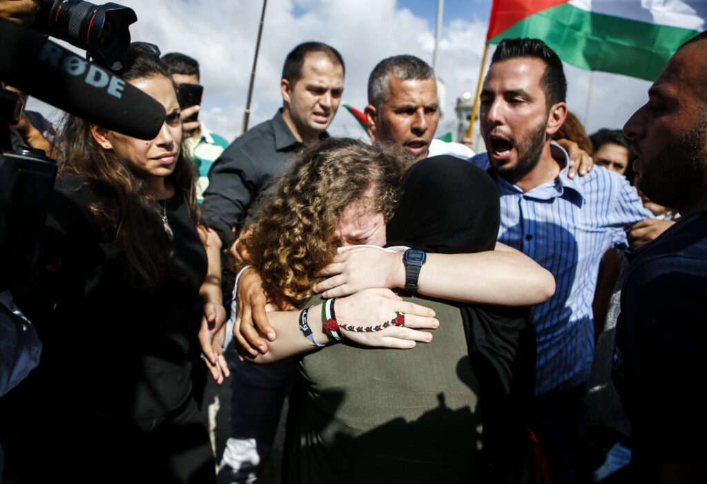 Two people—one visibly crying—hug amid a circle of people who cheer along. A camera and boom microphone are on the left side of the frame.