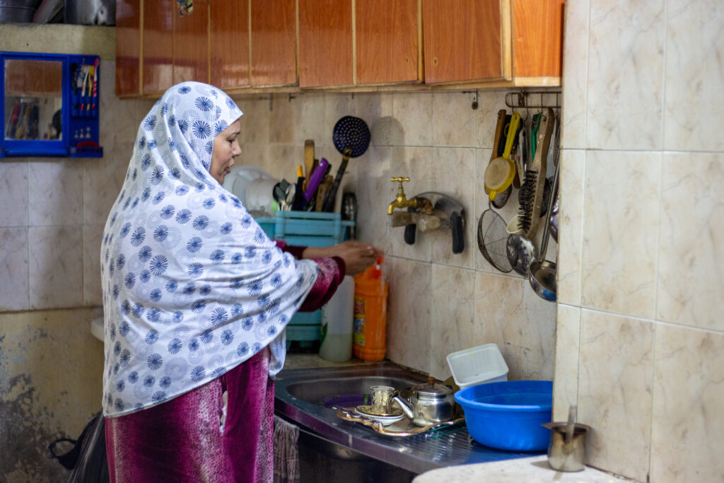 A woman in a blue and white scarf and dark pink dress washes dishes at a metal sink.