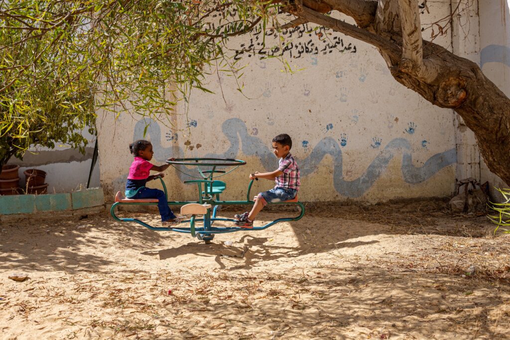 Two children sit on a turquoise play structure in a yard under a tree.