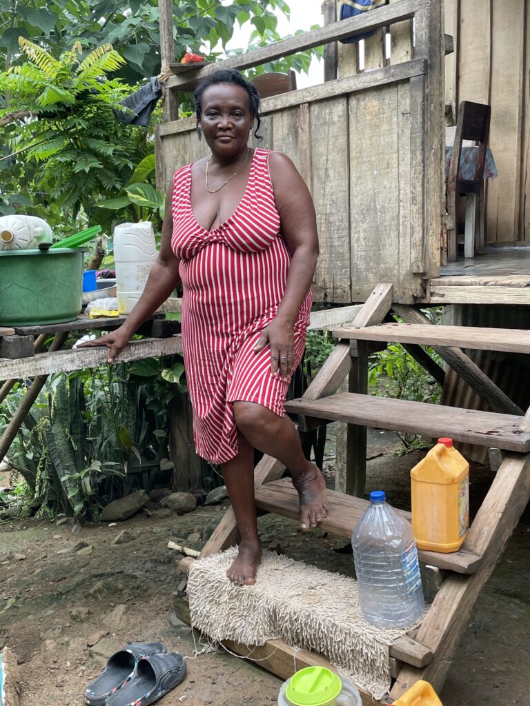A person with their hair in two braids wearing a red-and-white striped, knee-length dress stands on the front steps of a wooden house. Large jugs line the stairs, and pails sit on a table in the background.