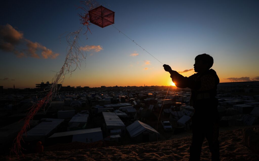 Against a bright orange sunset on the horizon, a silhouette of a child stands in the foreground with a village showing building rooftops in the background.