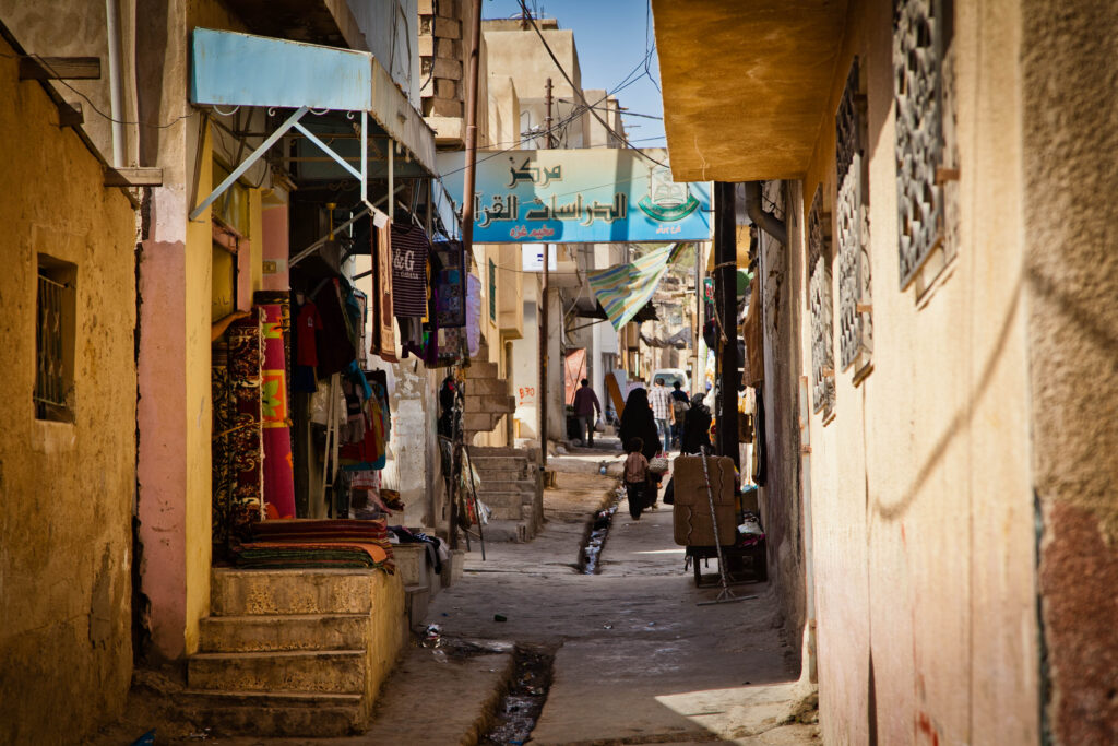 People walk down a narrow alleyway between tan concrete buildings.