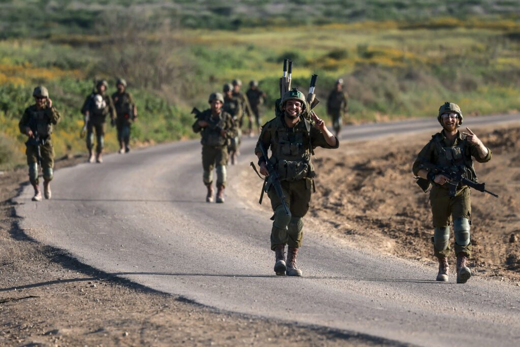 Several people wearing green uniforms, helmets, and boots carry guns and smile while walking on a paved road.