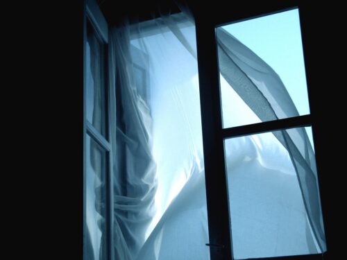 An open window separates a pitch-dark room on one side from an illuminated teal exterior and light blue curtains blowing in the breeze on the other.