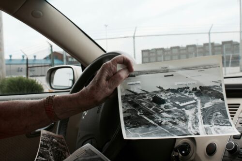 A person’s hand leans on a car’s steering wheel while holding a black-and-white aerial photograph of a section of town with particular sites such as Patapsco River, Dundalk, and Arundel Corp. Shipyard labeled.
