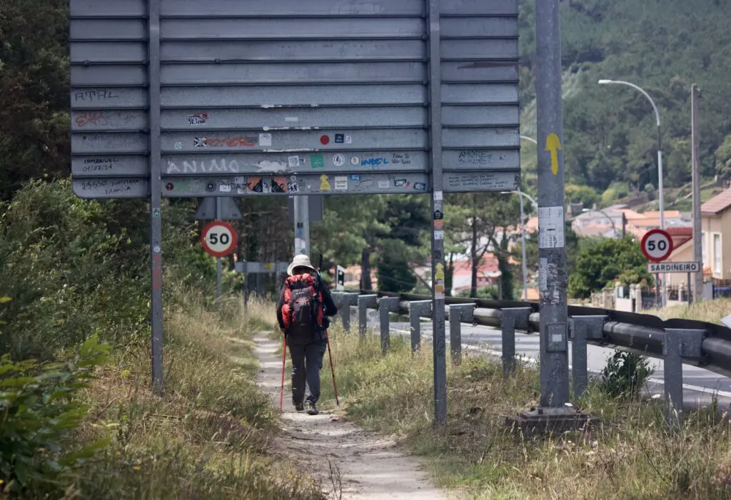 A person wearing a white hat, red-and-black backpack, and holding two hiking poles walks down a sandy trail lined with high grasses.