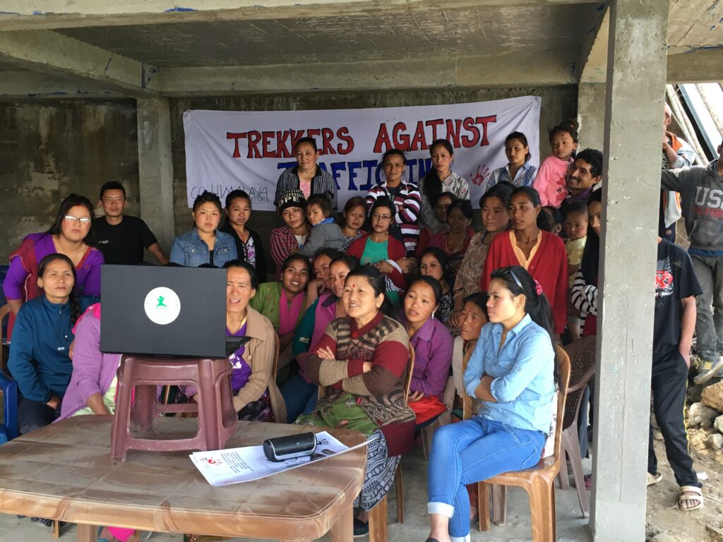 A large group of people—some seated and others standing behind them—gather under a stone roof and look at a black laptop sitting on a small maroon footstool placed on a wooden table.