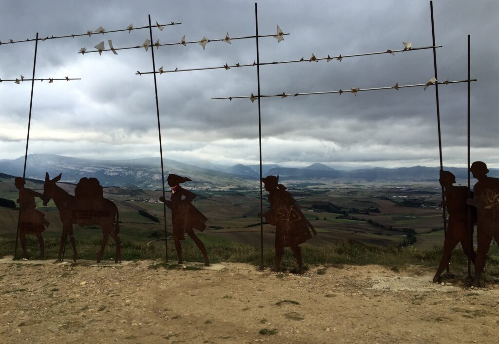 Metal sculptures that resemble silhouettes of people walking with a donkey are posted in a row along the edge of a dirt plot. Mountains line a horizon visible in the distance.