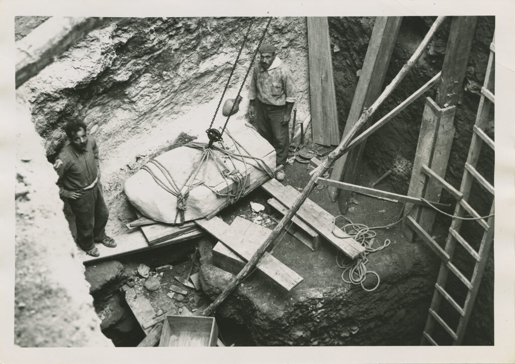 Inside a large excavation hole, two people stand on either side of a large block tied with rope that is being hoisted up by chains.