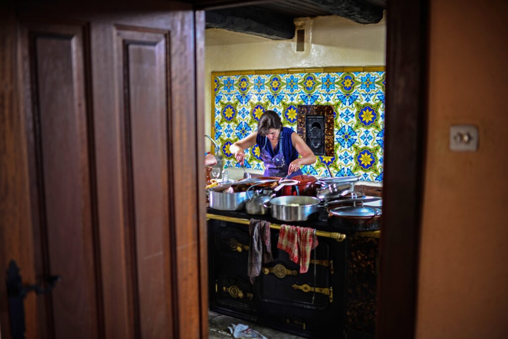 : A person wearing a blue apron stands behind and looks down at a stove, using utensils to stir contents in several steel pots and pans on top of it. A colorful mosaic fills the back wall.