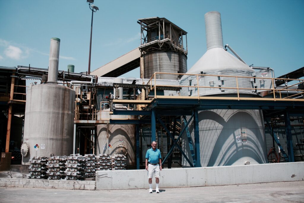 A person wearing short gray hair, a blue polo shirt, and tan shorts stands in front of a large factory building with metal silos and large holding tanks.