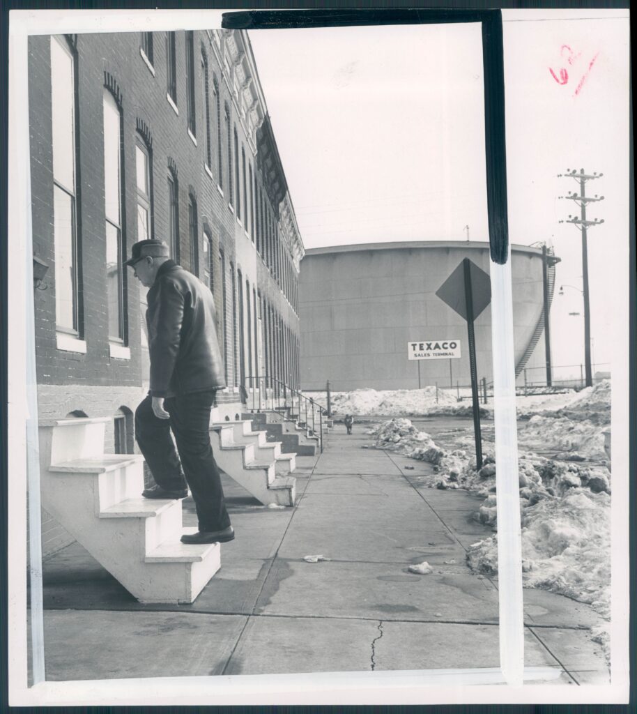 A black-and-white photograph shows a person wearing a cap, jacket, and slacks walking up steps into a brick building. A large container with a sign that reads “Texaco” on it is in the background.