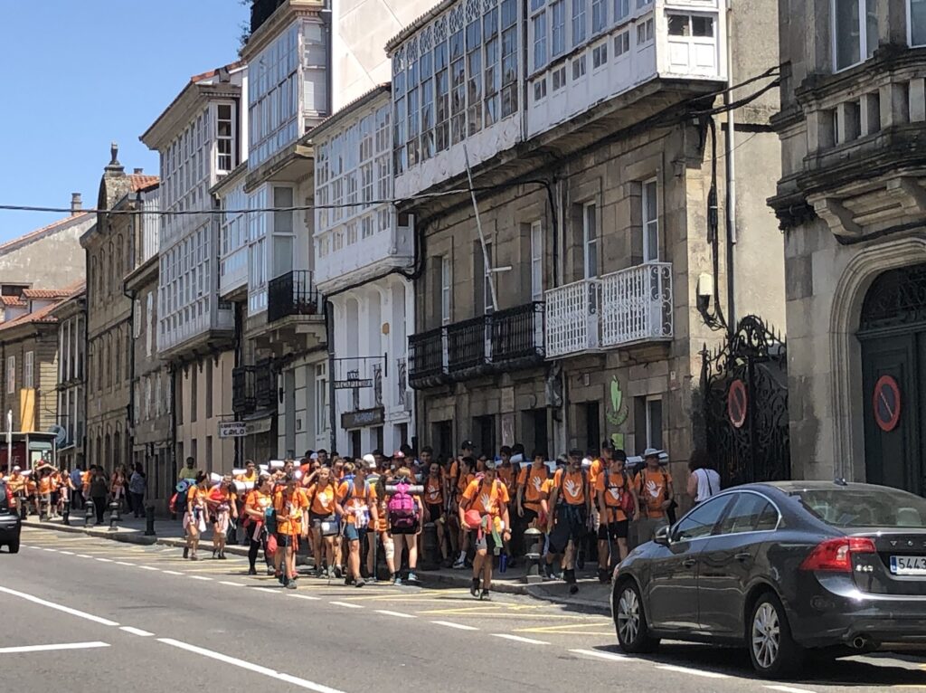 A large group of people in matching orange shirts with a white graphic on it walk down a narrow sidewalk between a row of stone buildings with balconies and a paved road. Some people overflow into the street.