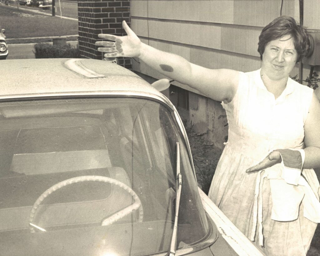 A person with short brown hair wearing a sleeveless dress gestures their arms toward a car. A streak is visible on the roof of the car, and dust covers the person’s palm.