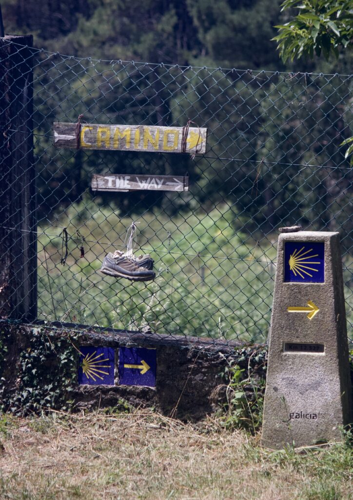 A pair of sneakers hang on a wire fence by the laces beneath wooden signs that read, “Camino” and “The way” with arrows pointing right. A stone object with a blue and yellow graphic, sitting to the right of the sneakers, has a yellow arrow pointing right as well.