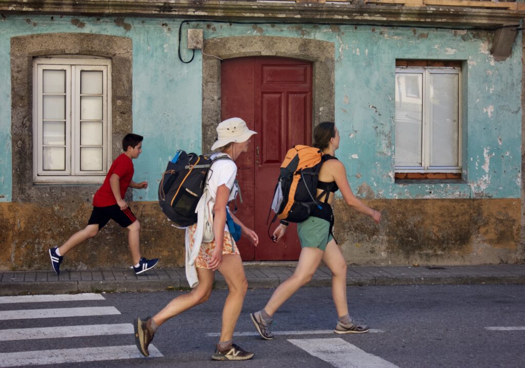Three people wearing shorts and sneakers—two of them wearing large backpacks—walk to the image’s right in the middle of a paved road. A building covered in weathering teal paint lines the background.