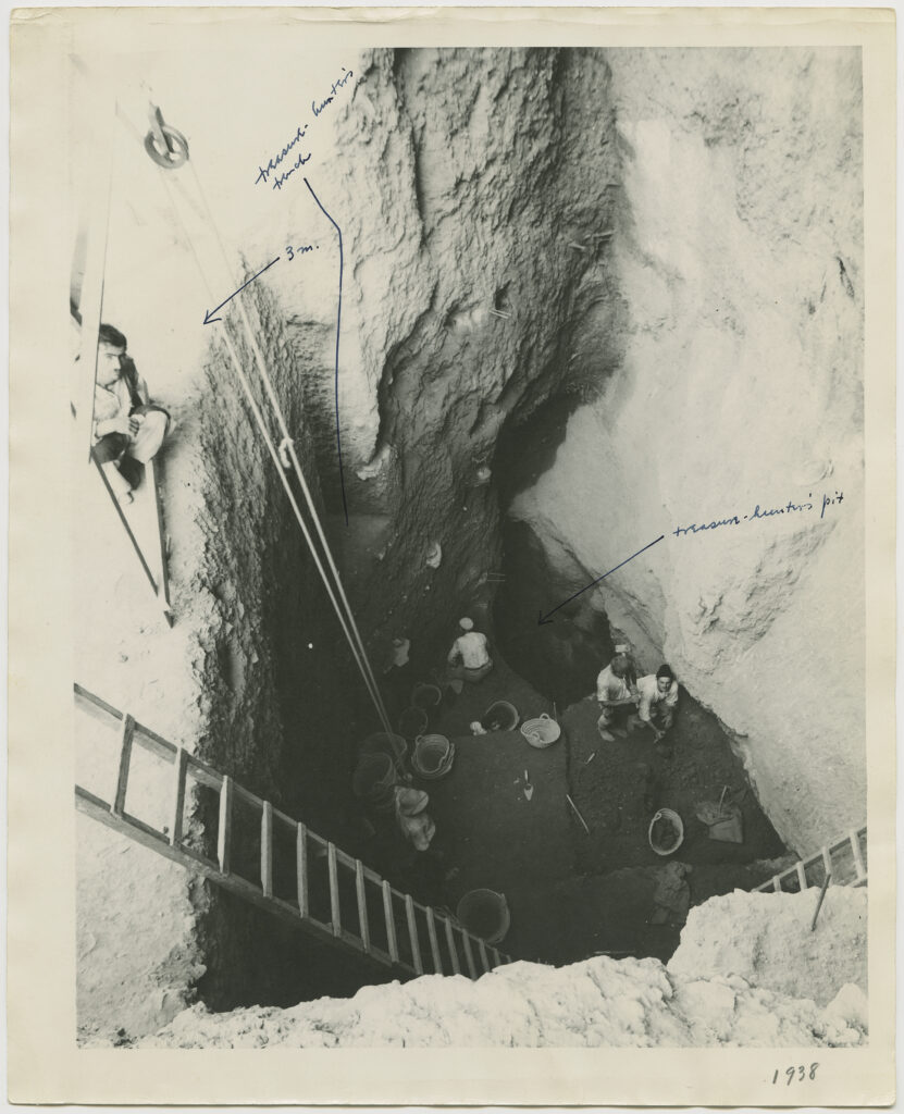 Shot from above, an image shows a ladder descending into a deep excavated hole in which people wearing helmets are standing. Surrounded by several trowels and buckets of dirt and rocks, one of them works while three look up at the camera.