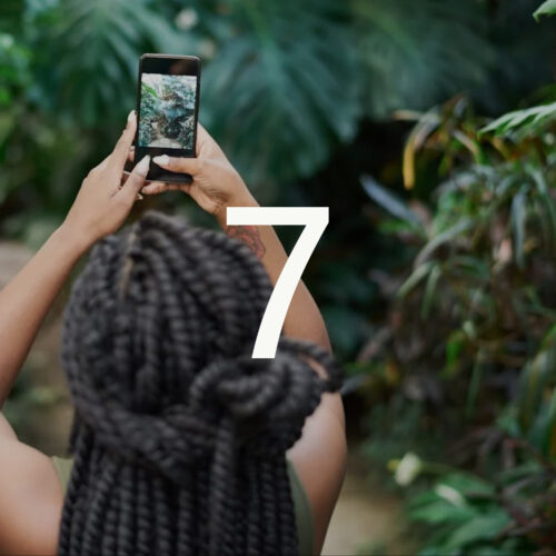 The image is a close-up on an individual from the back, focusing on their hands holding a smartphone with a nature picture on the screen. Large, stylized number "7" is partially visible, suggesting a series or season number. The background is a lush green blur, presumably foliage.