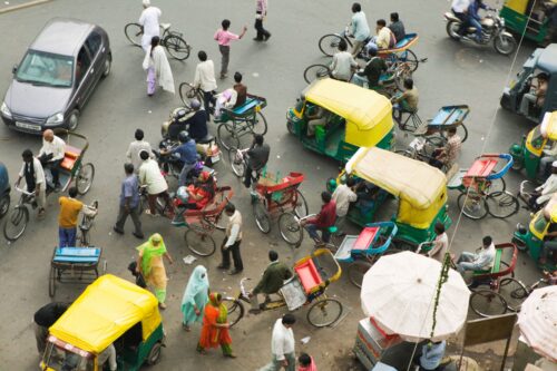 An overhead view shows a street intersection filled with a mix of pedestrians, bicycles, rickshaws, motorcycles, and cars facing in various directions.
