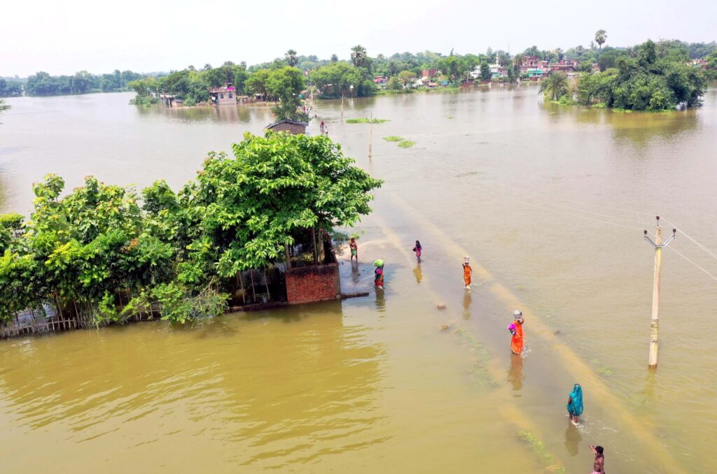 A line of people in colorful clothing wade through knee-deep floodwater on a submerged road surrounded by patches of land with trees and buildings.