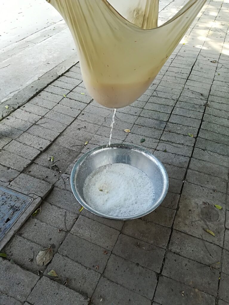 A tan cloth is used to strain frothy white soy milk into a metal bowl.