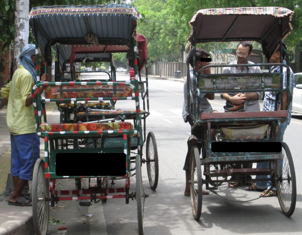 Two cycle rickshaws are parked on the side of a street; two drivers converse with a bearded person while another driver stands beside a rickshaw watching.