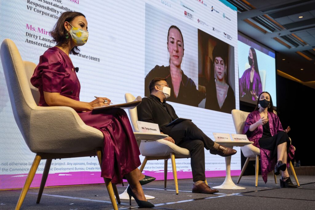 Three people wearing face masks sit in chairs on a stage in front of a screen showing images of two people.