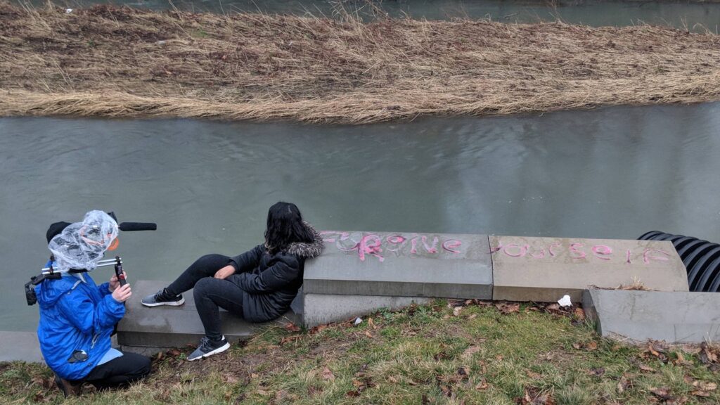 A person in a blue jacket holds a video camera next to a woman wearing a black jacket and black pants who sits on concrete slabs next to faded pink graffiti. Behind her is a gray river and beige grass on the riverbank.