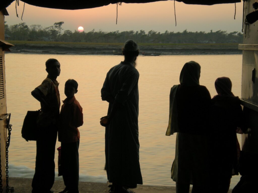 Silhouetted against a hazy sunset, a small group of people stand on a riverbank, looking out over the water.