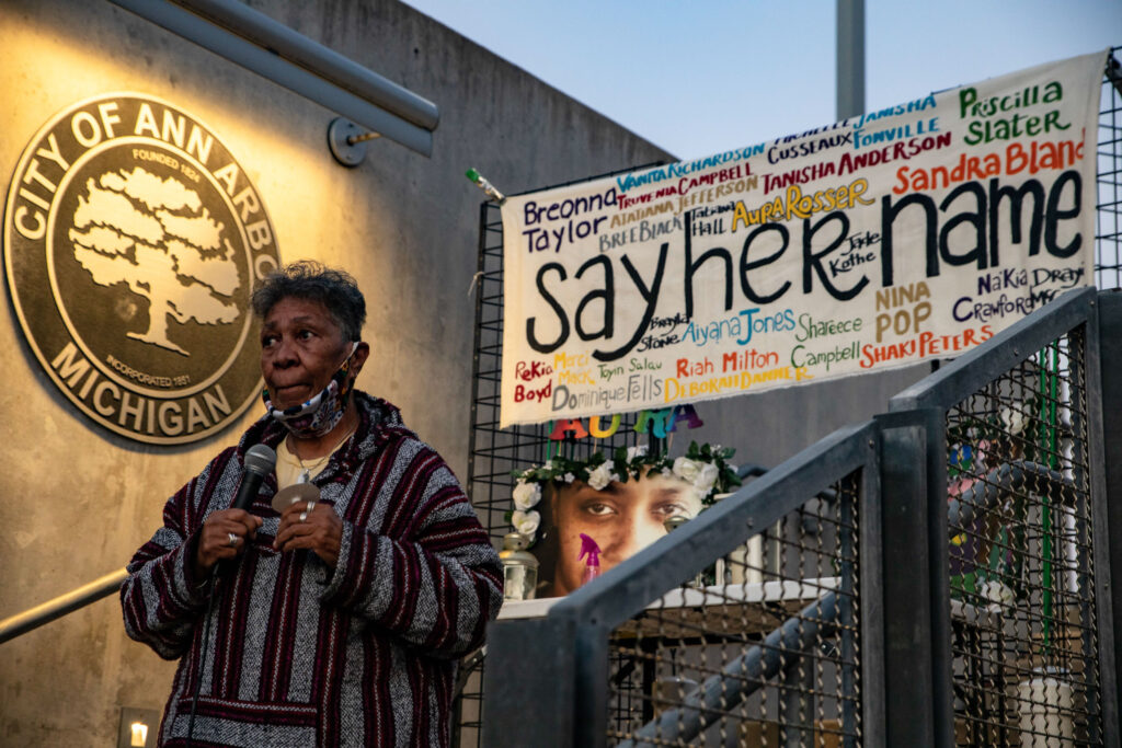 A woman with gray short hair stands on a metal staircase with a large circular plaque to her right that says, “City of Ann Arbor, Michigan.” To her left is a photo of a young woman’s face framed with white flowers and a large banner that reads, “say her name.” Various names are painted around the phrase, including but not limited to Breonna Taylor, Sandra Bland, Nina Pop, Priscilla Slater, Aura Rosser, Rekia Boyd, Shaki Peters, Toyin Salau, Atatianna Jefferson, and Tanisha Anderson.