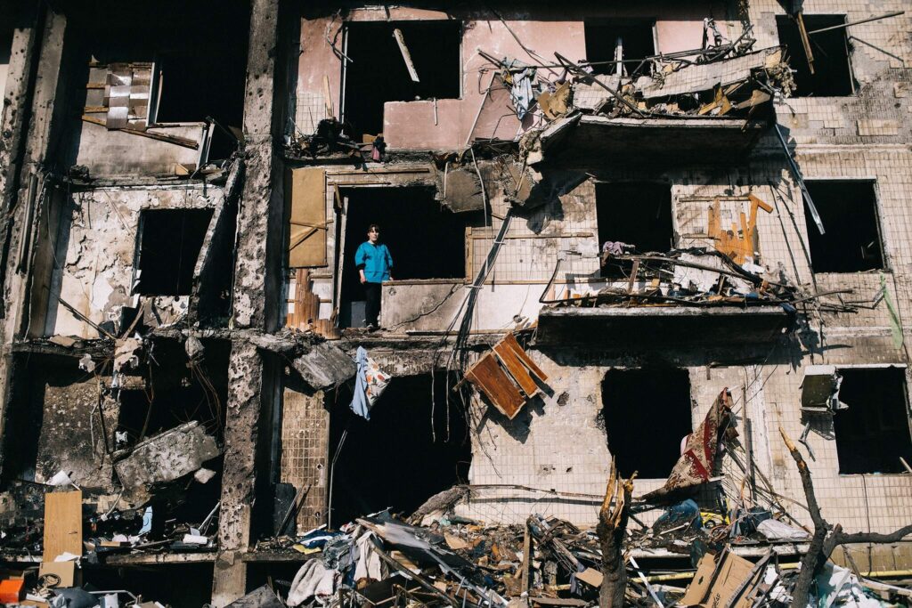 A person in a blue shirt stares out an opening on the side of a storied building where a window was. The structure has no windows left, smashed balconies, and burned and broken walls.