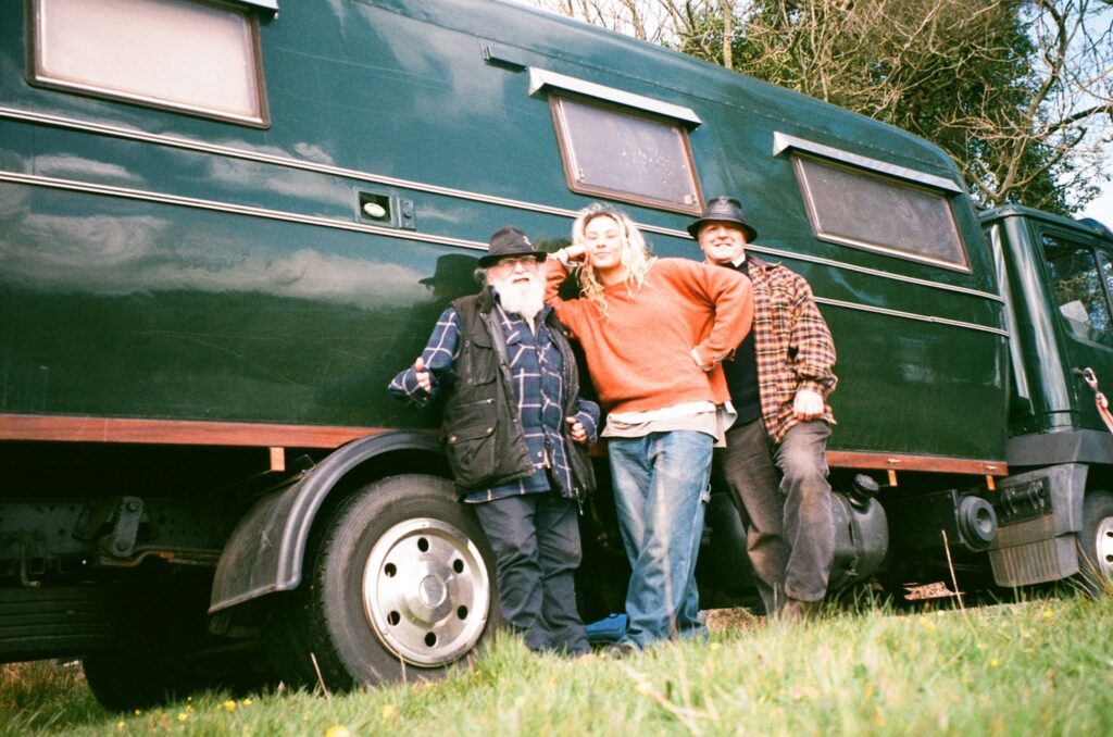 Three people wearing sweaters, flannel shirts, and jeans stand and pose in front of a green truck with an extended trailer attached.