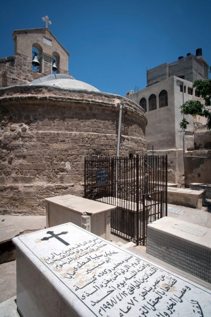 A white tombstone with a black cross and inscriptions stands in the foreground. In the background lies an old stone church holding a bell tower.