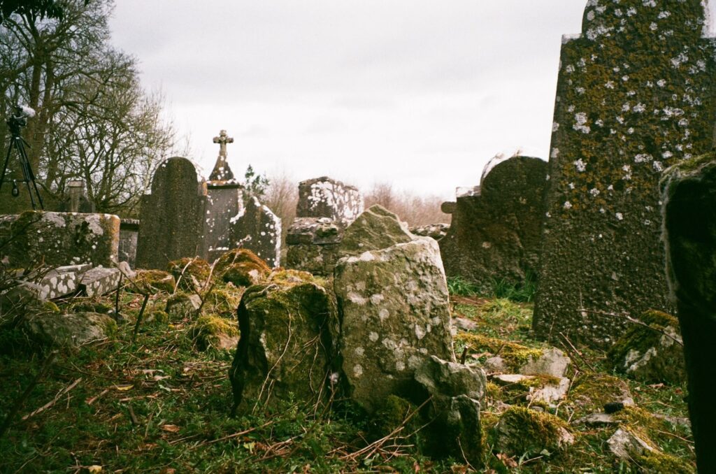 Rock and several tombstones, one with a cross on top, are scattered around a grassy plot of land. They are covered in moss, lichen, twigs, and vines.
