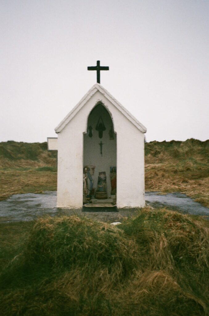 A small, white structure open in the front has a cross on top and stands in an open field.