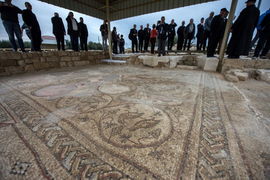 A group of people stands under a canopy, observing large, detailed ancient mosaics with intricate designs and patterns that were uncovered during an archaeological excavation.