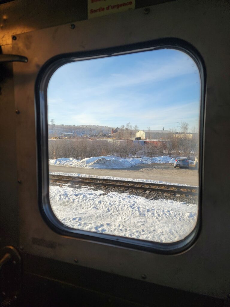 A view through a train window shows a snowy treed landscape with railroad tracks in the foreground.