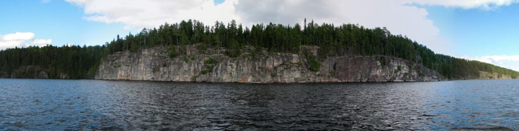 A panoramic photograph features a forested rocky shoreline from across a calm lake. The cliffs are lined with dense green trees under a partly cloudy sky.